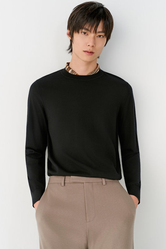 A man wearing a black long-sleeve crewneck sweater and beige trousers, standing against a light gray background.