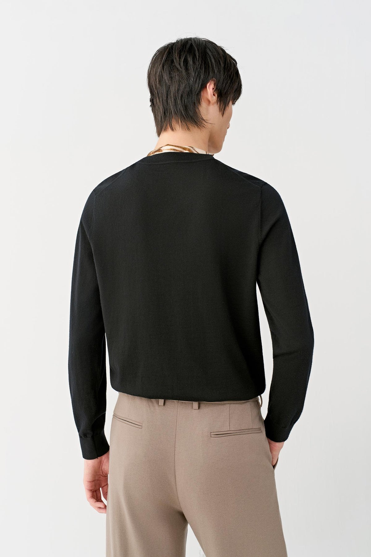 A man shown from the back wearing a black long-sleeve knit sweater and beige trousers, photographed against a clean white studio background.