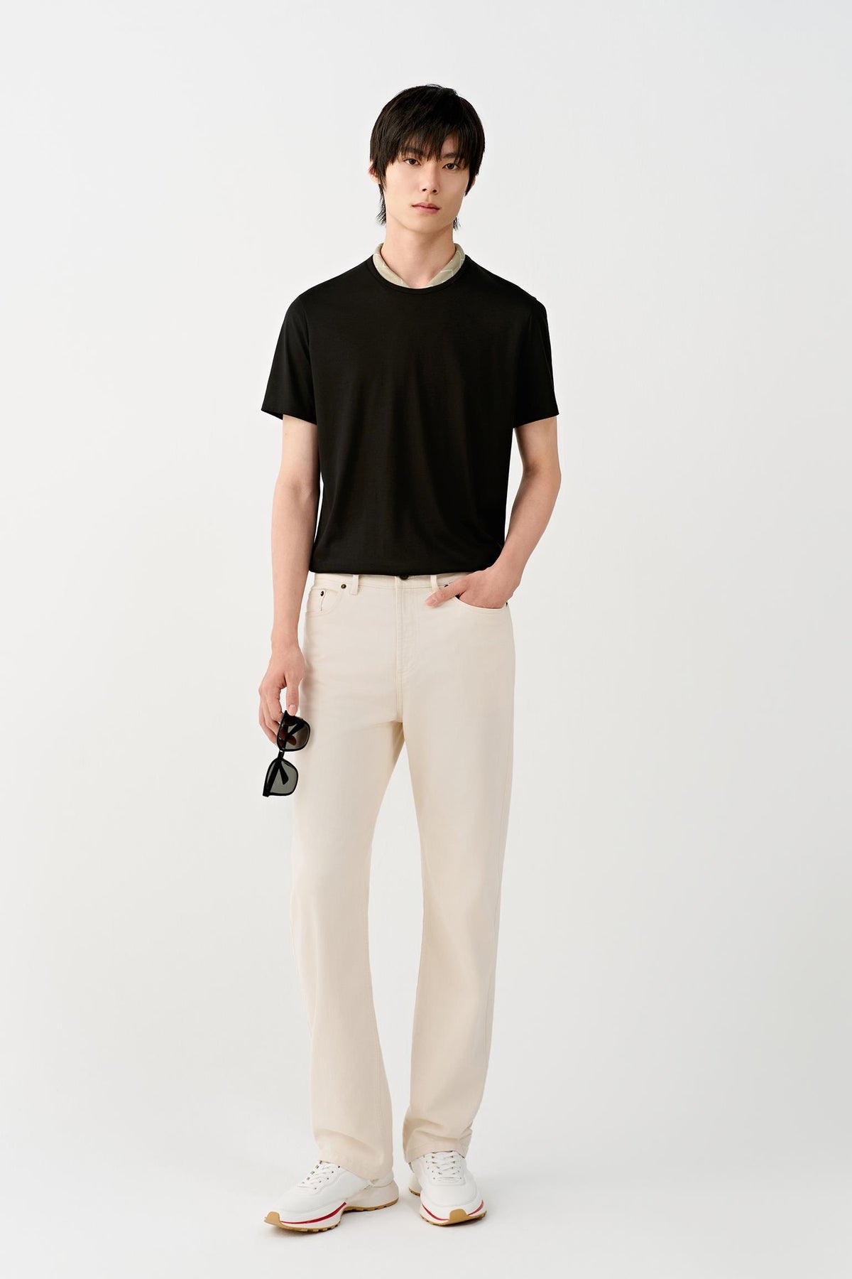 A male model stands against a plain light gray studio background wearing a black short-sleeve crew-neck t-shirt, off-white straight-leg trousers, and white sneakers, holding a pair of sunglasses in one hand.