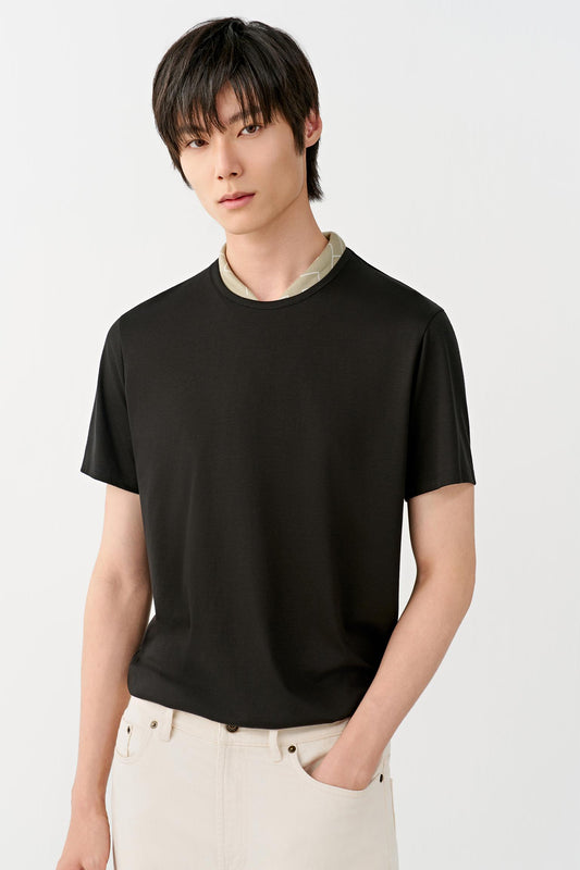 A man wearing a black short-sleeve crewneck t-shirt with a light beige collar insert, paired with off-white trousers, against a plain light gray background.