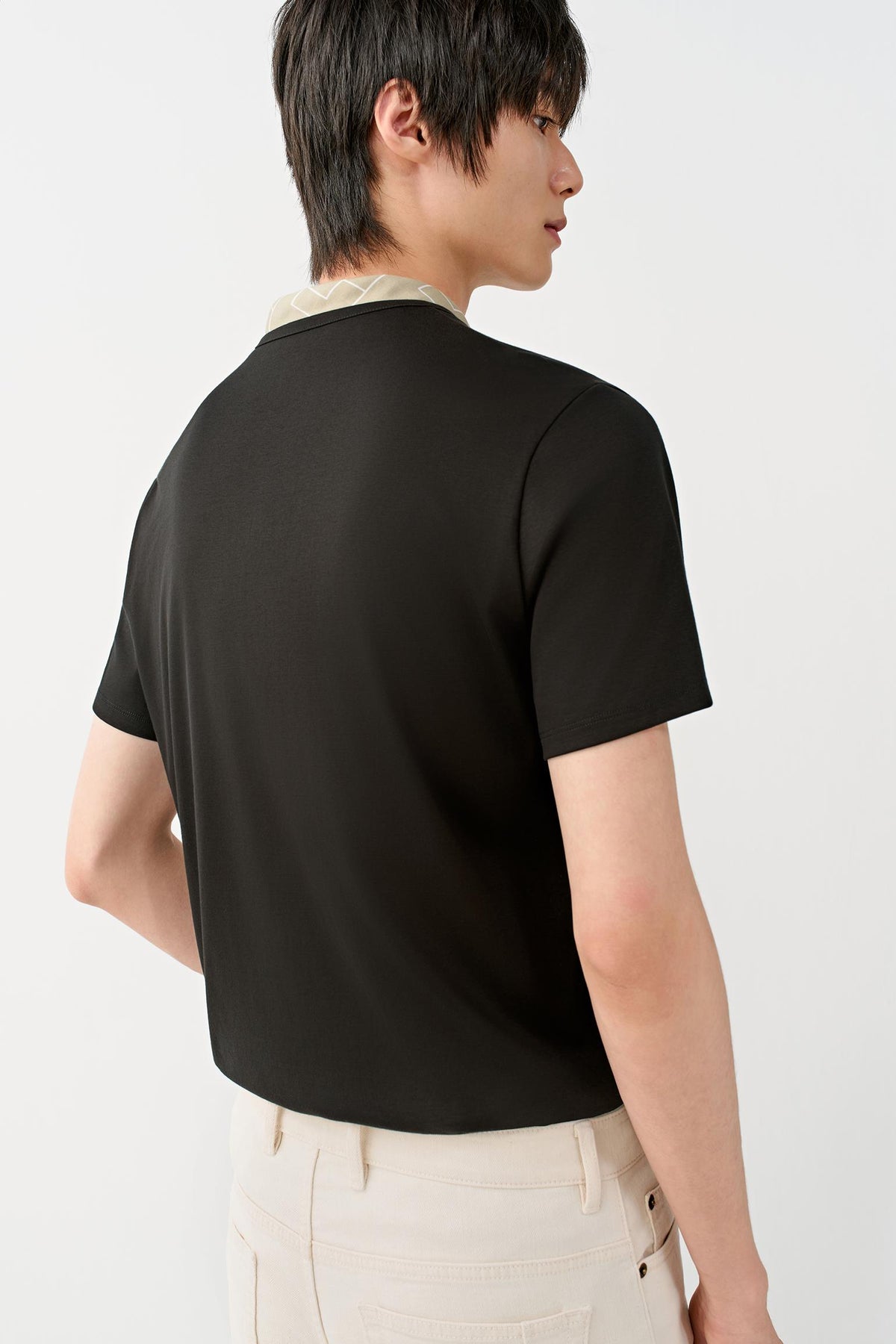 A man shown from the back wearing a black short-sleeve crewneck t-shirt with a light beige collar detail, paired with off-white trousers, against a plain white studio background.