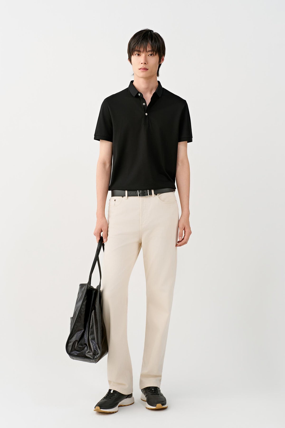 A man wearing a black short-sleeve polo shirt, cream-colored trousers, and a black belt, with a black shoulder bag, photographed against a clean white studio background.