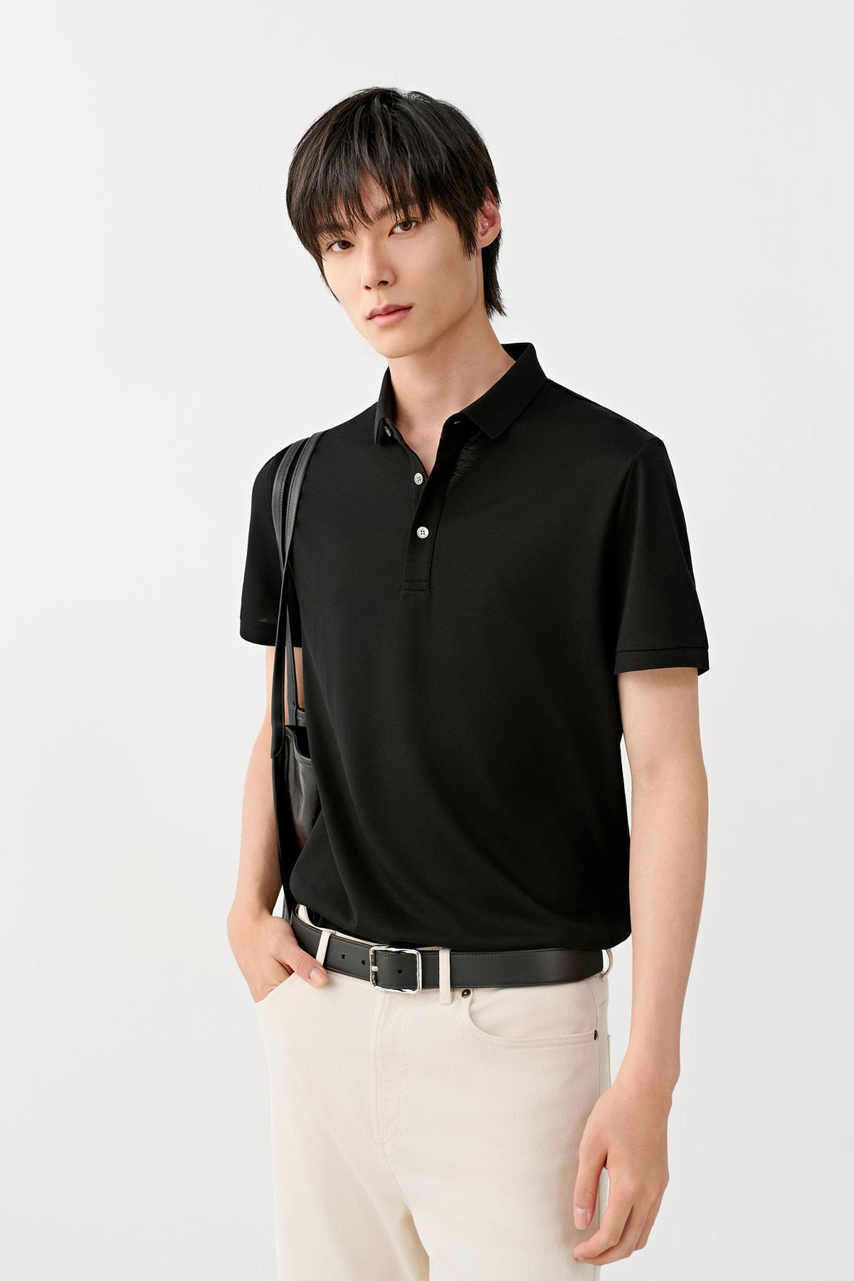 A man wearing a black short-sleeve polo shirt, cream-colored trousers, and a black belt, with a black shoulder bag, photographed against a clean white studio background.