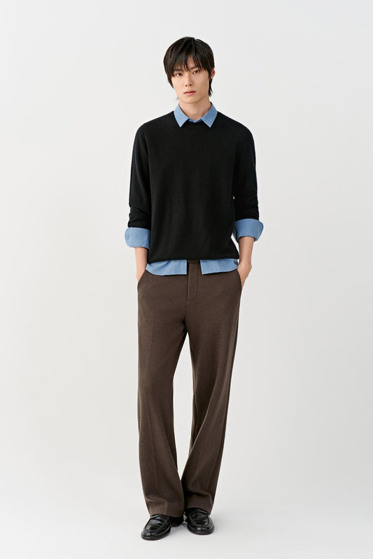A man modeling a black long-sleeve knit sweater layered over a light blue collared shirt, paired with brown trousers and black shoes, against a plain light gray studio background.
