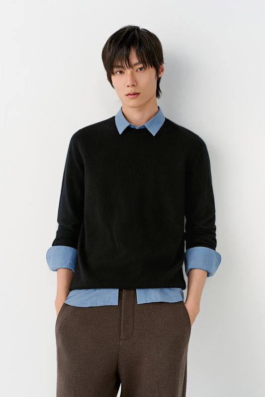 A man wearing a black knit sweater layered over a light blue collared shirt, paired with brown trousers, standing against a plain light gray wall.