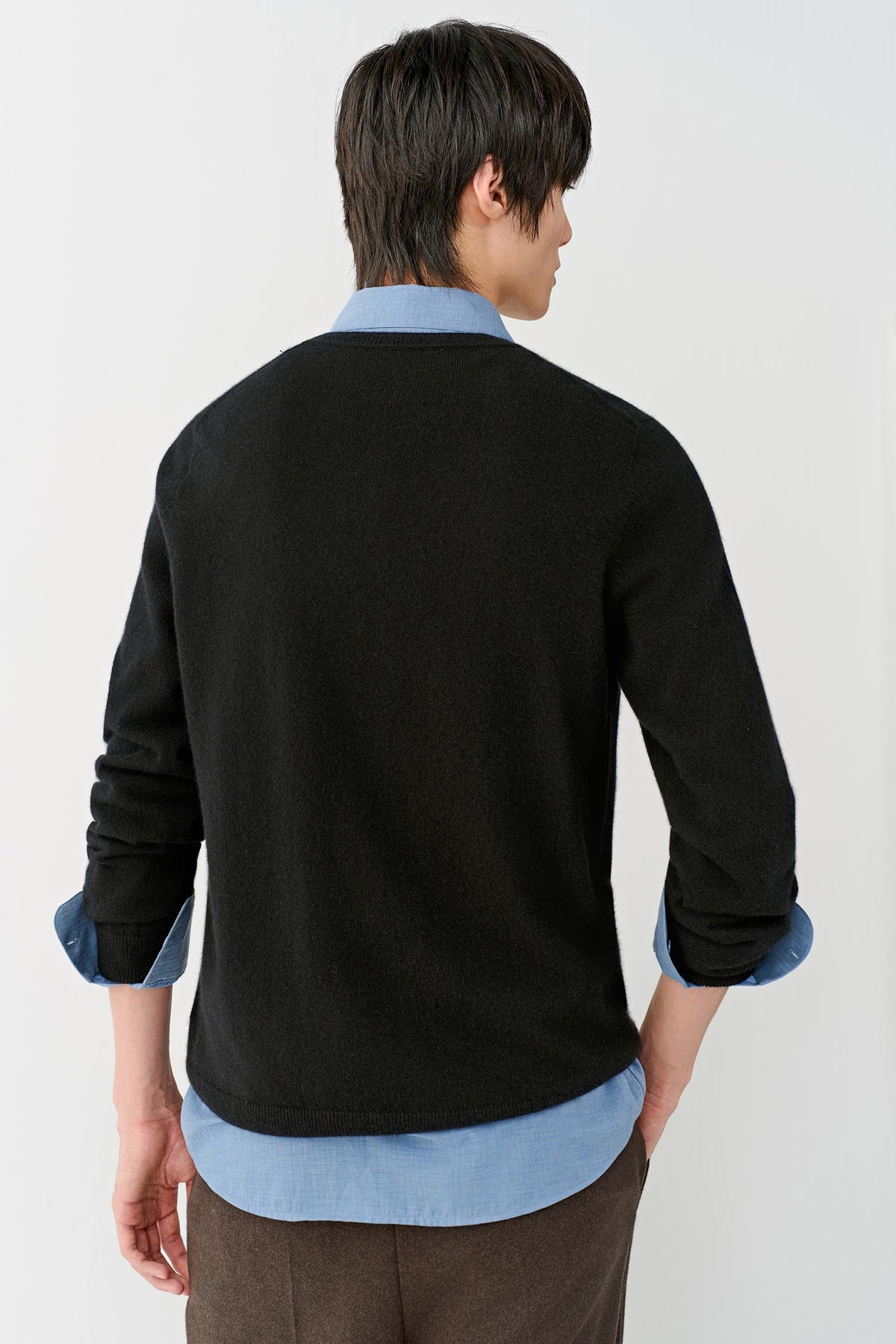A man shown from the back wearing a black knit cardigan over a light blue collared shirt, paired with brown trousers, against a plain light gray studio background.