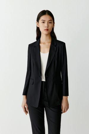 Full-length studio photograph of a woman wearing a deep black blazer in worsted wool. The blazer has a straight tailored silhouette, notched lapels, a single-breasted front and long sleeves. It is worn open over a white top and matching black tailored trousers. The jacket falls below the hips and creates a clean elongated line.