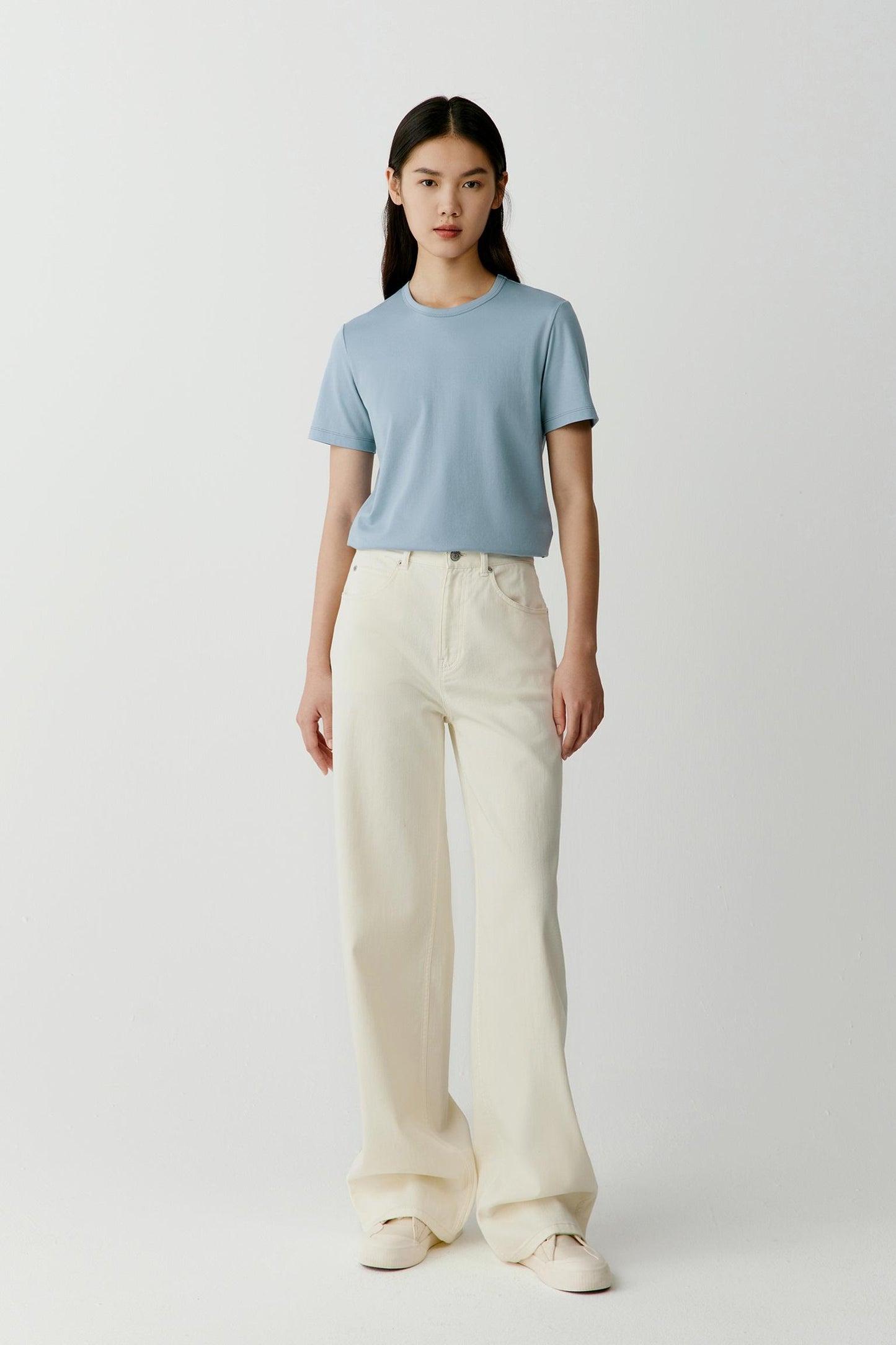 Full-length front view showing the T-shirt styled with off-white trousers. The straight cut, round neckline and short sleeves create balanced proportions, while the lightweight cotton jersey drapes softly and follows the body with ease.
