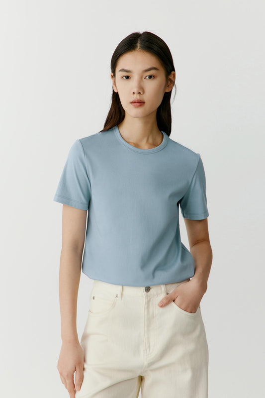 Front view of a woman wearing a light blue cotton jersey T-shirt, highlighting the smooth jersey surface, soft hand and lightweight construction. The fresh blue shade emphasizes the clean simplicity of the design, while the fabric falls naturally to create a relaxed and fluid silhouette.
