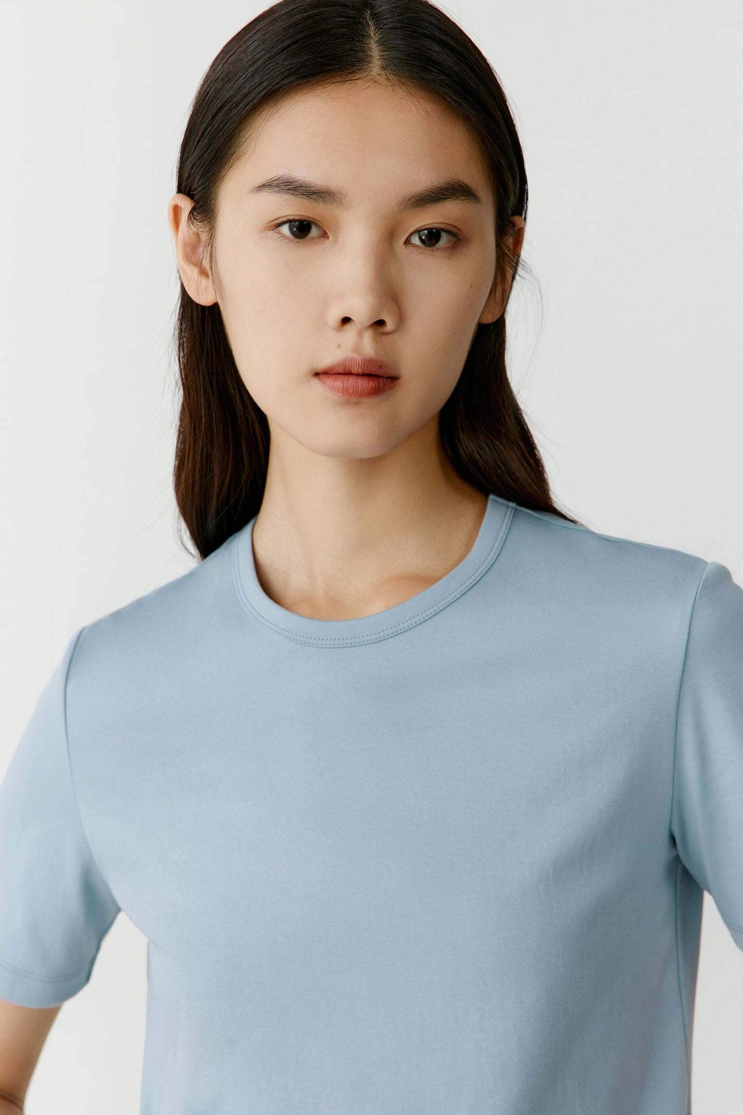 Close-up portrait focusing on the round neckline, shoulder construction and upper body. The image highlights the smooth matte surface, even jersey knit and soft texture, while showcasing the subtle contrast between the light blue shade and the clean lines of the garment.