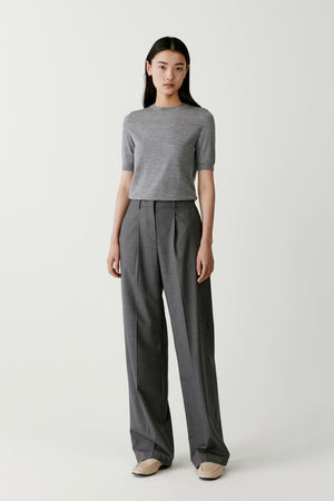 Full-length front view of a model wearing a grey short-sleeved tee knitted from superfine wool. The tee features a straight relaxed silhouette with a classic round neckline and short sleeves that fall midway along the upper arm. The fine knit structure creates a smooth and lightweight surface with a soft matte finish. The hem falls naturally at hip level and the garment is styled with wide grey trousers.