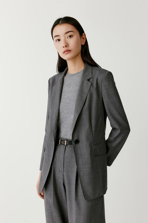Full-length studio photograph of a woman wearing a light grey blazer in worsted wool. The blazer has a straight tailored silhouette with a lightly defined waist, notched lapels, a single-breasted front and long straight sleeves. It is worn open over a matching grey top and matching tailored trousers. The model stands facing forward against a light grey studio background with one hand placed behind her back.