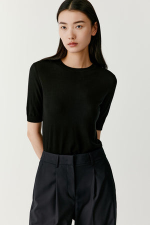 Front view of a model wearing a black short-sleeved tee knitted from superfine machine-washable wool. The tee features a straight relaxed silhouette with a classic round neckline and short sleeves extending halfway down the upper arm. The lightweight knit creates a smooth and breathable surface.