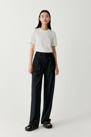 Full-length front view of a model wearing a white T-shirt made from organic cotton jersey. The T-shirt has a relaxed straight silhouette with a classic round crew neckline and short sleeves. The lightweight jersey fabric falls softly over the torso. It is paired with high-waisted black wide-leg trousers and flat black shoes.