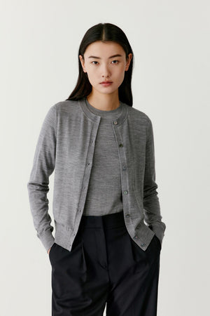 Front view of a model wearing a grey wool cardigan with a relaxed straight silhouette. The cardigan features a classic round neckline and a central button placket with tonal buttons. The long sleeves extend to the wrists and the soft wool knit creates a smooth, slightly textured surface. The cardigan falls naturally over the torso and is paired with dark trousers.