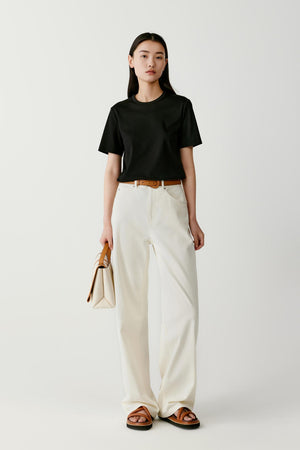 Full-length front view of a model wearing a black short-sleeved T-shirt made from cotton jersey. The T-shirt has a relaxed straight silhouette with a classic round crew neckline and short sleeves. The lightweight jersey fabric drapes softly over the torso. It is styled with high-waisted off-white wide-leg trousers and flat brown sandals, with a small beige handbag held in one hand.