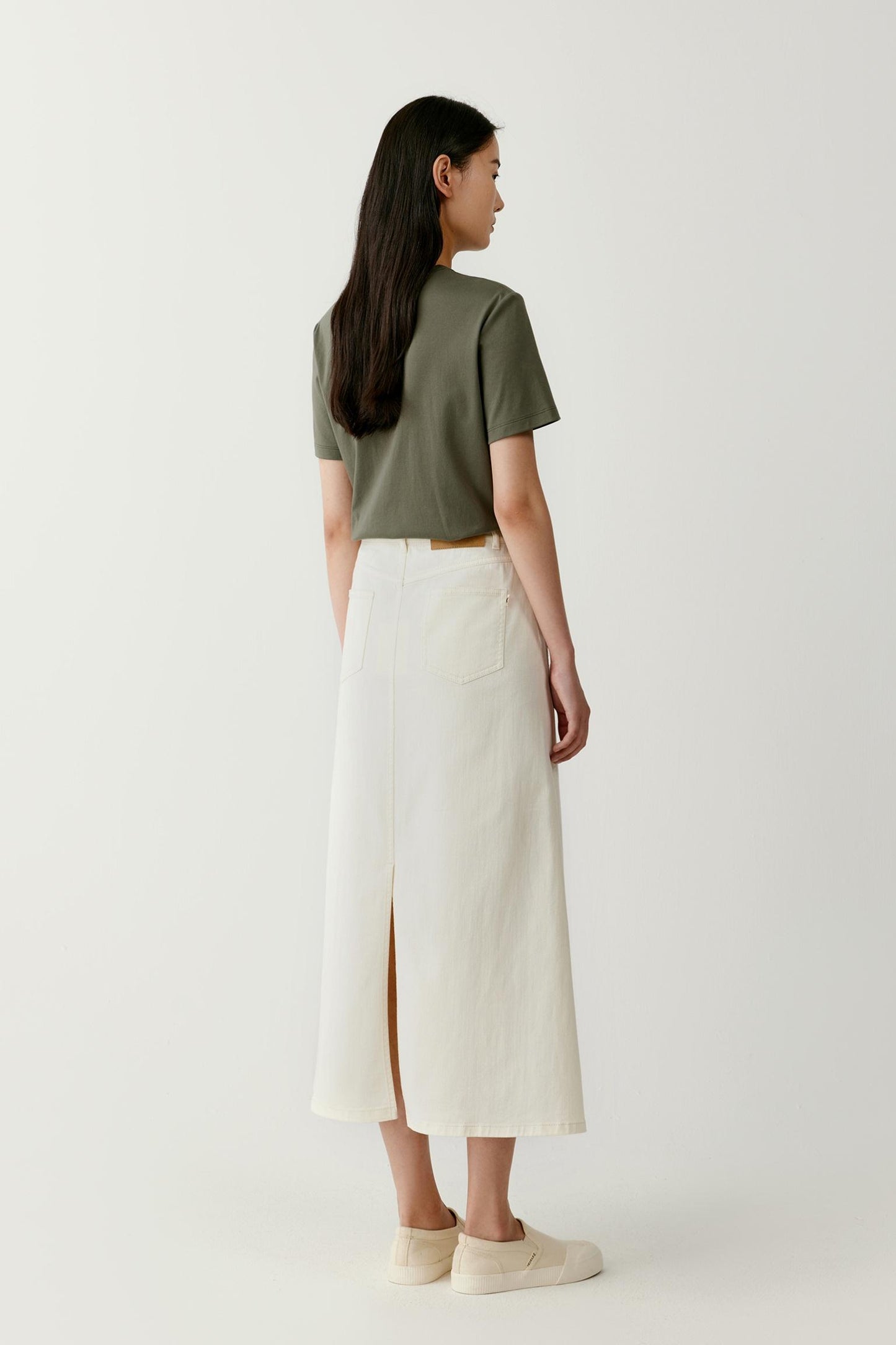 Full-length back view presenting the T-shirt styled with a long light-coloured skirt. The image highlights the balanced proportions, straight cut and lightweight structure of the jersey fabric, demonstrating how the garment integrates into a relaxed everyday silhouette.