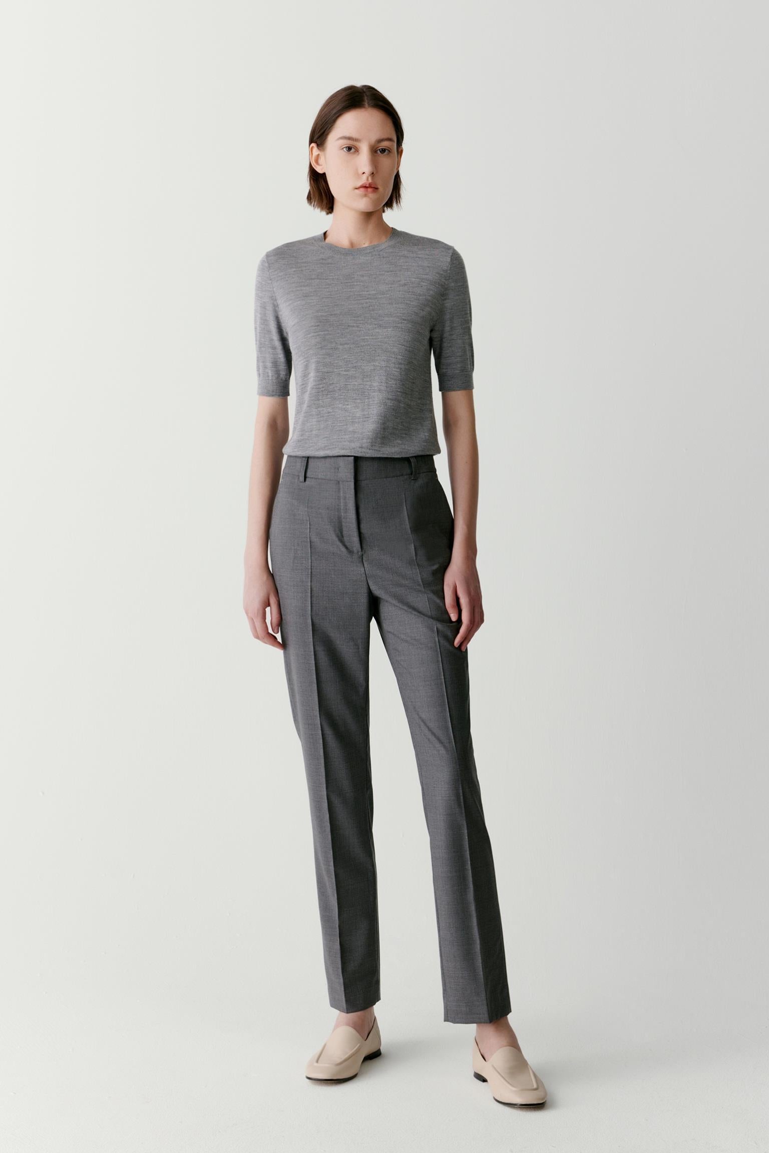 Front view of a woman wearing grey tapered trousers in worsted wool, highlighting the clean silhouette, softly structured drape and refined appearance of the fabric. The tapered leg and streamlined proportions create a balanced, tailored look.