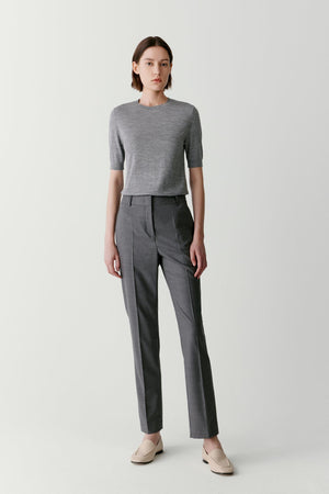 Front view of a woman wearing grey tapered trousers in worsted wool, highlighting the clean silhouette, softly structured drape and refined appearance of the fabric. The tapered leg and streamlined proportions create a balanced, tailored look.