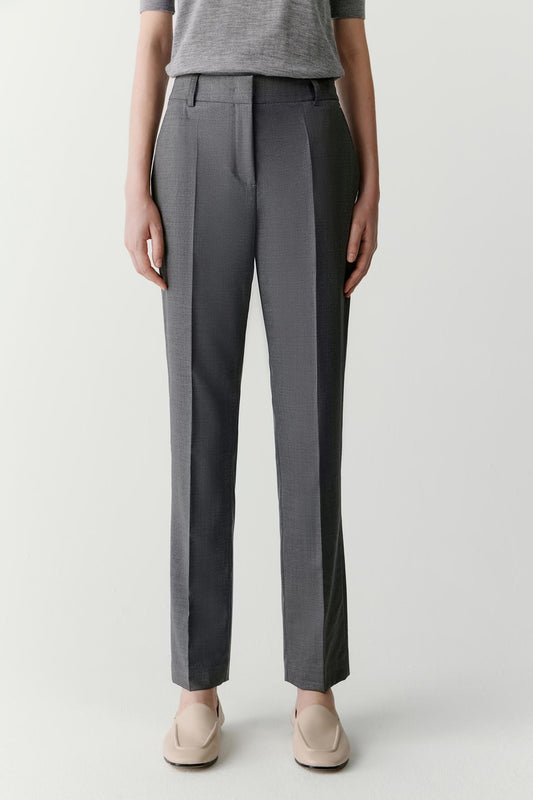 Full-length view focused on the trousers, showing the tapered cut from hip to ankle, the smooth surface of the worsted wool and the precise construction of the leg. The fabric falls cleanly while maintaining subtle structure.