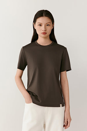 Front view of a woman wearing a dark grey cotton jersey T-shirt, highlighting the smooth jersey surface, soft hand and lightweight construction. The clean design and deep neutral shade emphasize the simplicity of the silhouette, while the fabric falls naturally with a fluid drape.