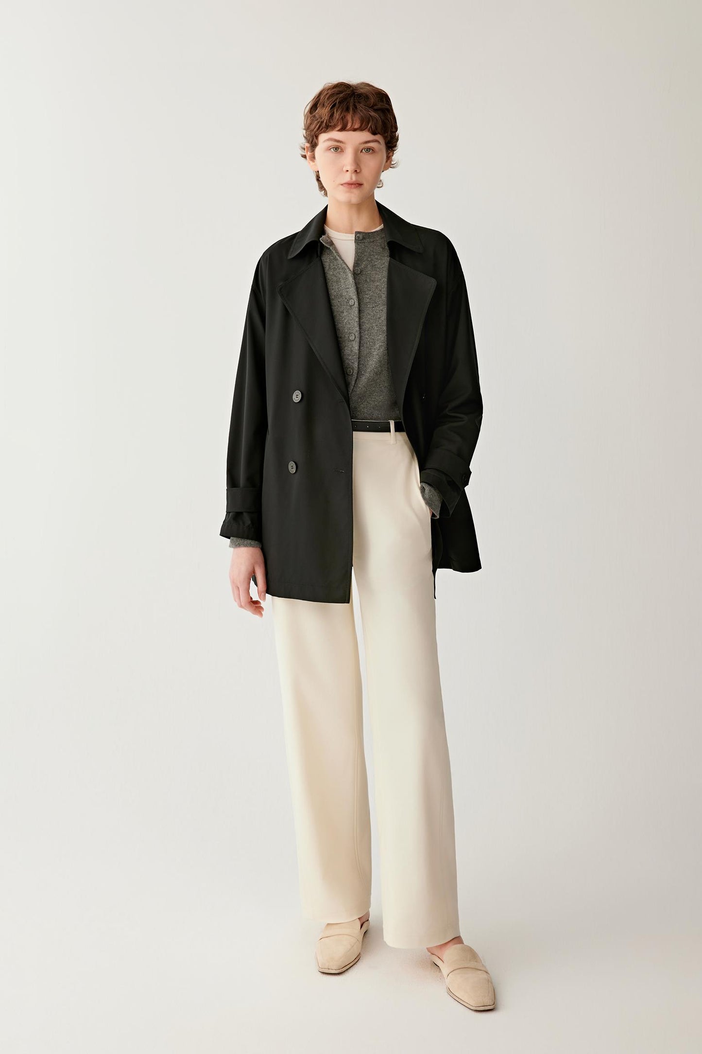 Full-length front view showing the proportions of the short trench coat. The image emphasizes the softly structured shape, fluid movement of the fabric and the clean lines created by the tailored construction and belted waist.