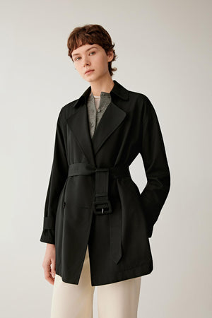 Front view of a woman wearing a short trench coat in deep black wool and silk. The image highlights the clean, structured silhouette, soft drape and smooth surface of the fabric. A double-breasted front, notched lapels and a self-fabric belt define the waist, creating a balanced and tailored appearance.