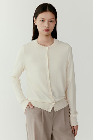 Front view of a model wearing an ivory wool cardigan with a refined straight silhouette. The cardigan features a round neckline, a central button placket with tonal buttons and long sleeves extending to the wrists. The fine wool knit creates a smooth, soft surface.
