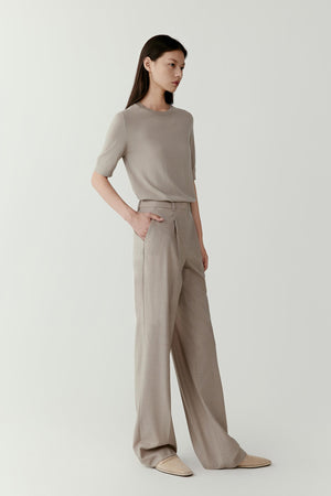 Front view of a model wearing straight-leg trousers crafted from worsted wool in a light neutral shade. The trousers feature a fluid, elongated silhouette with clean lines that fall naturally from the waist to the hem. The worsted wool fabric has a smooth surface and refined structure, creating a balanced and elegant appearance.