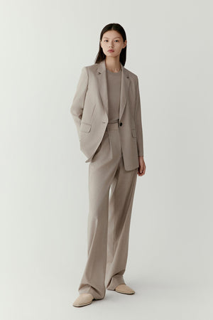 A woman stands wearing a light taupe melange blazer in worsted wool with matching trousers, captured against a neutral backdrop.