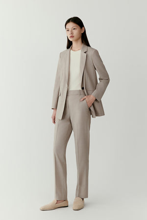 Front view of a model wearing tapered trousers crafted from worsted wool in a soft neutral tone. The trousers feature a clean, tailored silhouette with a fitted waist and gently tapered leg that narrows toward the ankle. The worsted wool fabric provides a smooth surface and lightweight structure.