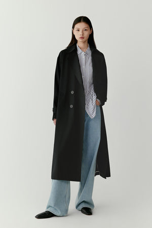 Full-length studio photograph of a woman wearing a long deep black trench coat in wool and silk with a minimalist straight silhouette and single-breasted construction. The coat falls below the calf and is styled with wide light blue trousers.
