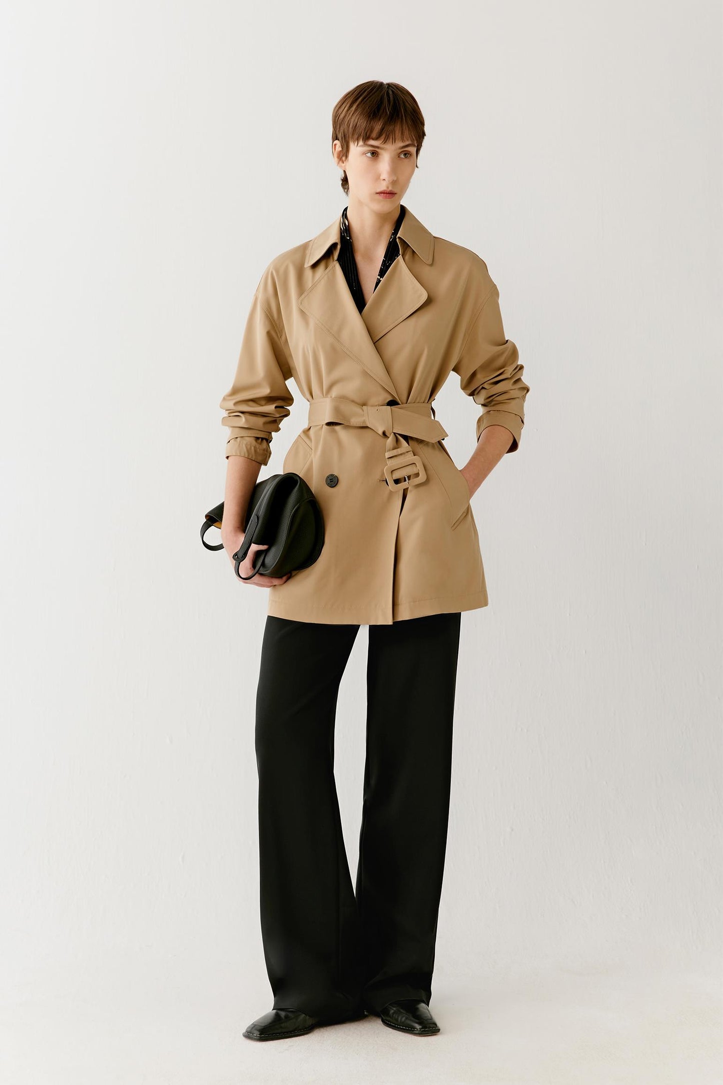 Front view of a woman wearing a short trench coat in a camel shade crafted from wool and silk. The image highlights the clean, structured silhouette, soft drape and smooth fabric surface. A double-breasted front, notched lapels and a self-fabric belt define the waist, creating balanced proportions and a tailored appearance.