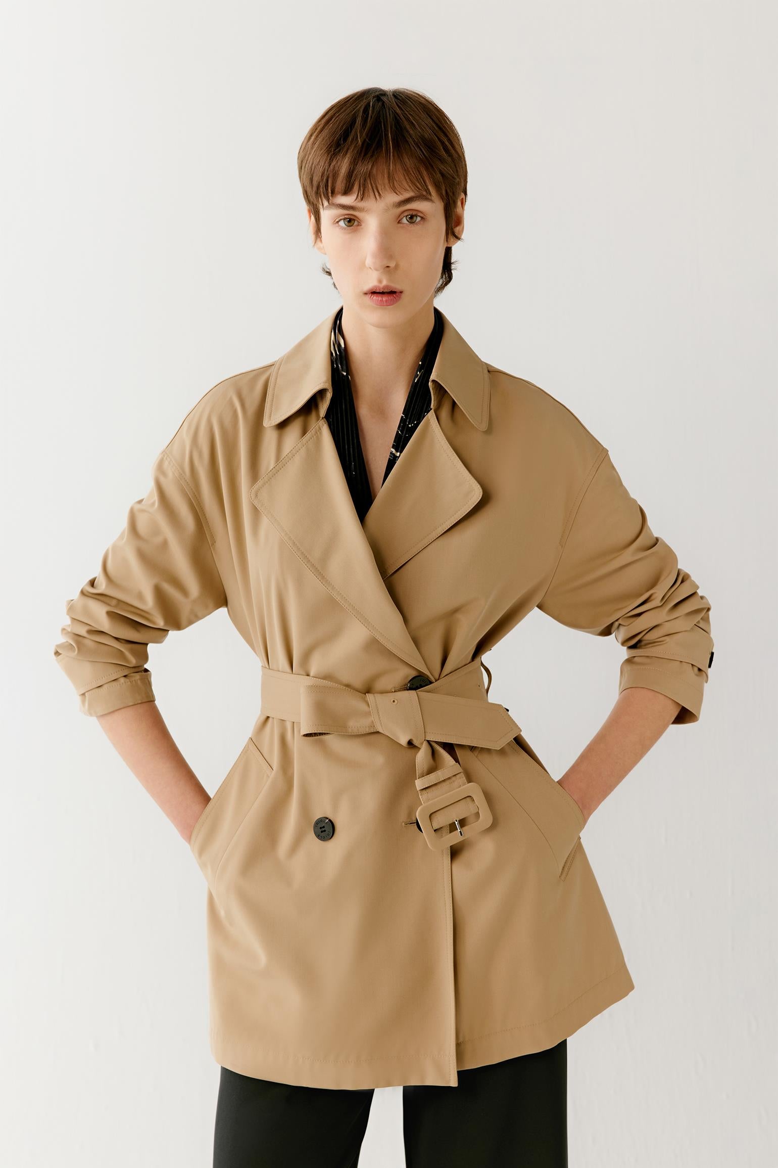 Three-quarter view showing the full silhouette and belted construction of the trench coat. The image emphasizes the fluid drape of the fabric, the softly structured shape and the clean lines that define the garment. The camel shade enhances the subtle depth and refined character of the design.