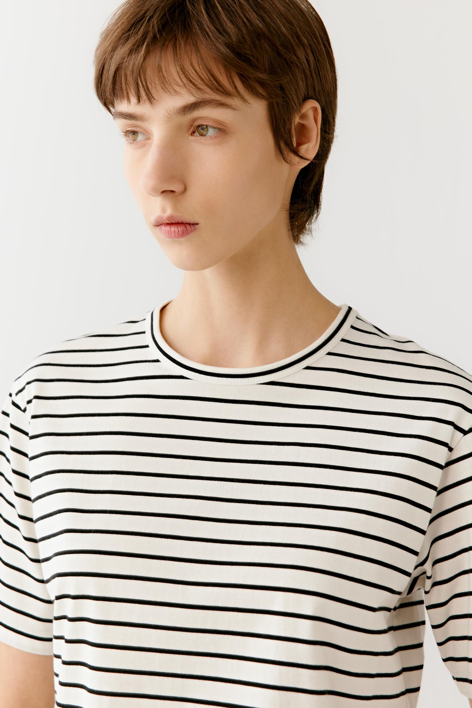 Three-quarter view showing the T-shirt styled with relaxed grey trousers. The lightweight jersey drapes naturally from the shoulders, while the round neckline, short sleeves and straight cut create a simple, structured silhouette with ease of movement.