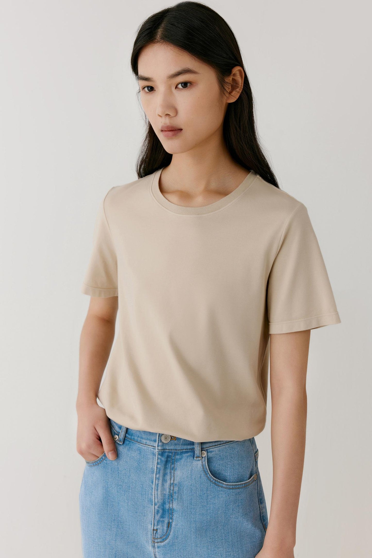 Front view of a woman wearing a beige T-shirt in cotton jersey. The image highlights the smooth surface, soft hand and lightweight structure of the fabric. The relaxed silhouette falls naturally through the body, while the round neckline and short sleeves create a clean and versatile appearance.