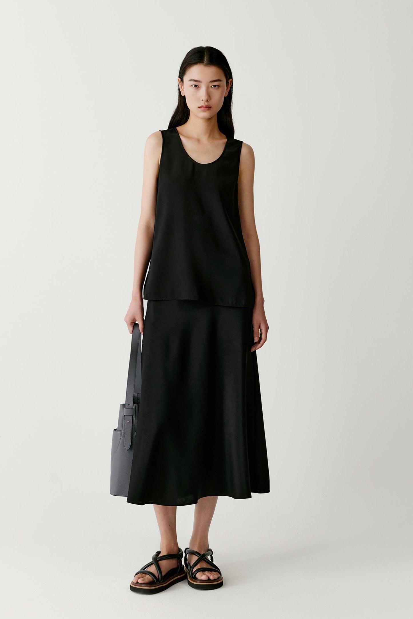 Front view of a woman wearing a sleeveless top in black silk crêpe de Chine. The image highlights the fluid drape, smooth surface and soft hand of the fabric. The relaxed silhouette falls naturally from the shoulders, while the round neckline and sleeveless construction create a clean and elongated appearance.