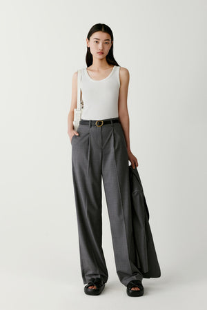 Front view of a model wearing straight-leg trousers crafted from natural worsted wool in a soft grey shade. The trousers feature a clean, elongated silhouette that falls evenly from the waist to the hem. The worsted wool fabric has a smooth surface and refined structure, allowing the garment to maintain crisp lines while draping naturally along the legs. The trousers are styled with a sleeveless white top and black shoes.