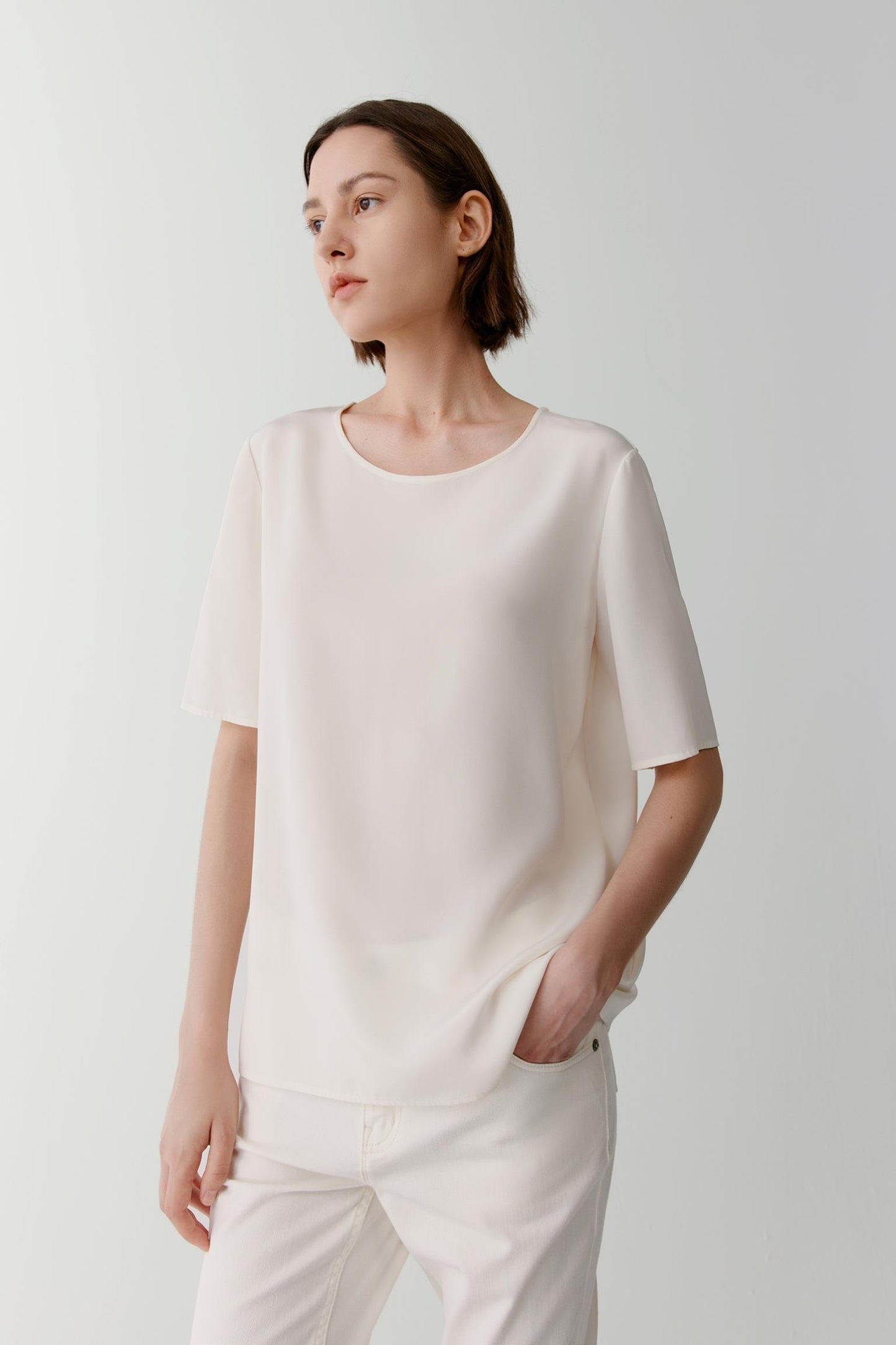 Front view of a woman wearing a shirt in white silk crêpe de Chine. The image highlights the fluid drape, smooth surface and soft hand of the fabric. The relaxed silhouette falls naturally from the shoulders, while the round neckline and short sleeves create a clean, understated appearance.