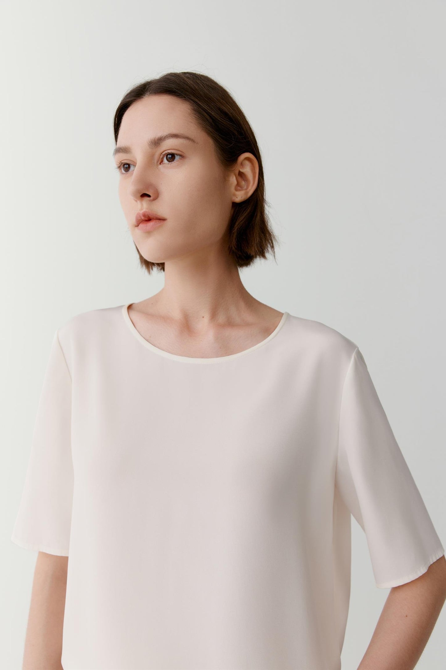 Close-up portrait focusing on the round neckline, shoulder line and upper construction. The image reveals the smooth texture, subtle lustre and refined finish of the silk crêpe de Chine fabric while showcasing the clean lines of the design.