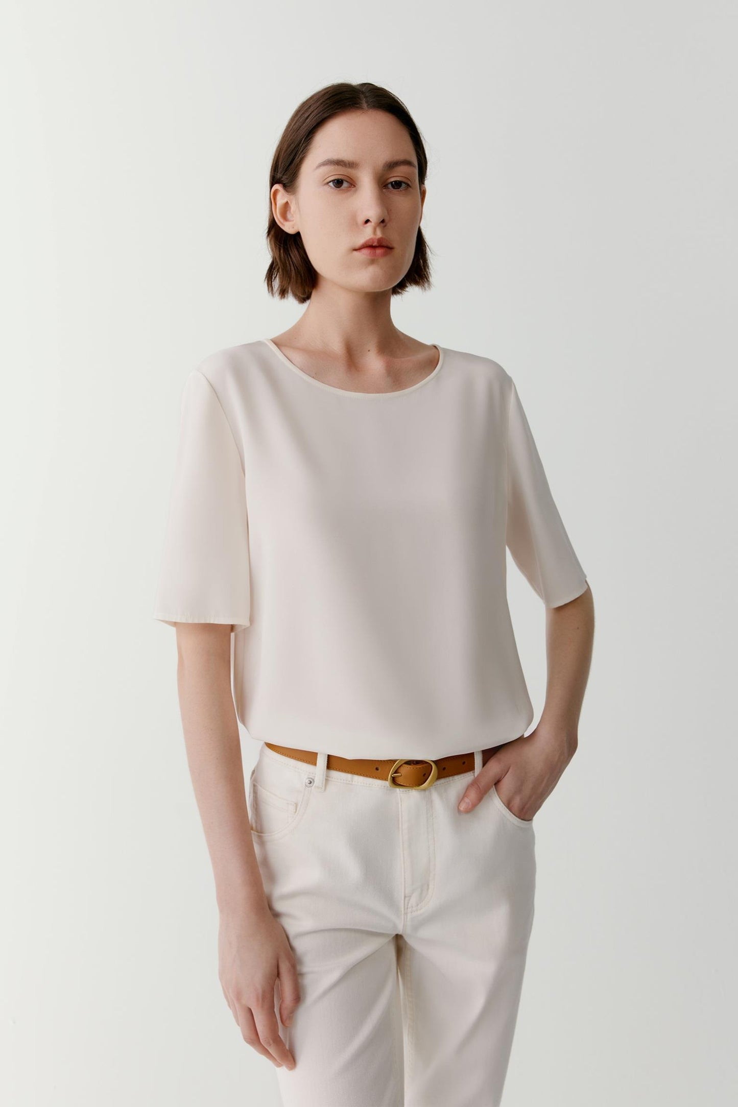 Close-up front view of a woman wearing a shirt in white silk crêpe de Chine. The image focuses on the round neckline, short sleeves and relaxed silhouette. The smooth fabric surface reveals a soft hand and fluid drape, while the clean construction and subtle lustre of the silk crêpe de Chine create a refined, understated appearance. The lightweight material falls softly over the body, highlighting the simplicity and balance of the design.
