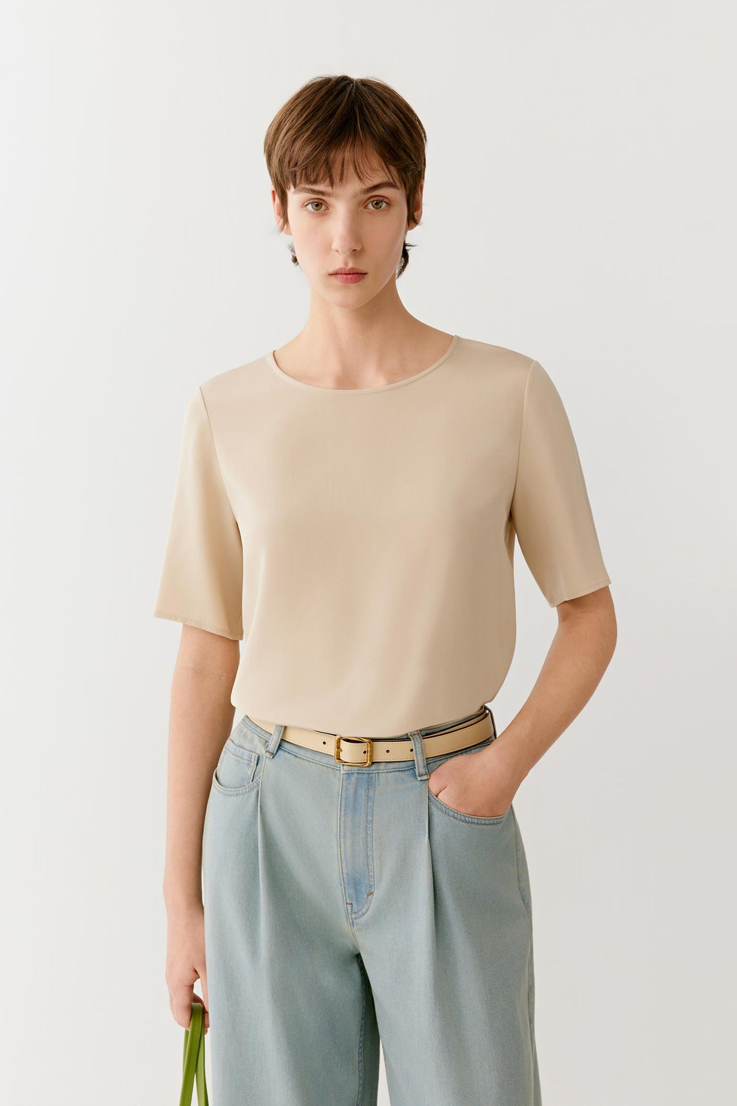 Front view of a woman wearing a shirt in light beige silk crêpe de Chine. The image highlights the fluid drape, smooth surface and soft hand of the fabric. The relaxed silhouette falls naturally from the shoulders, while the wide round neckline and short sleeves create a clean and understated appearance.