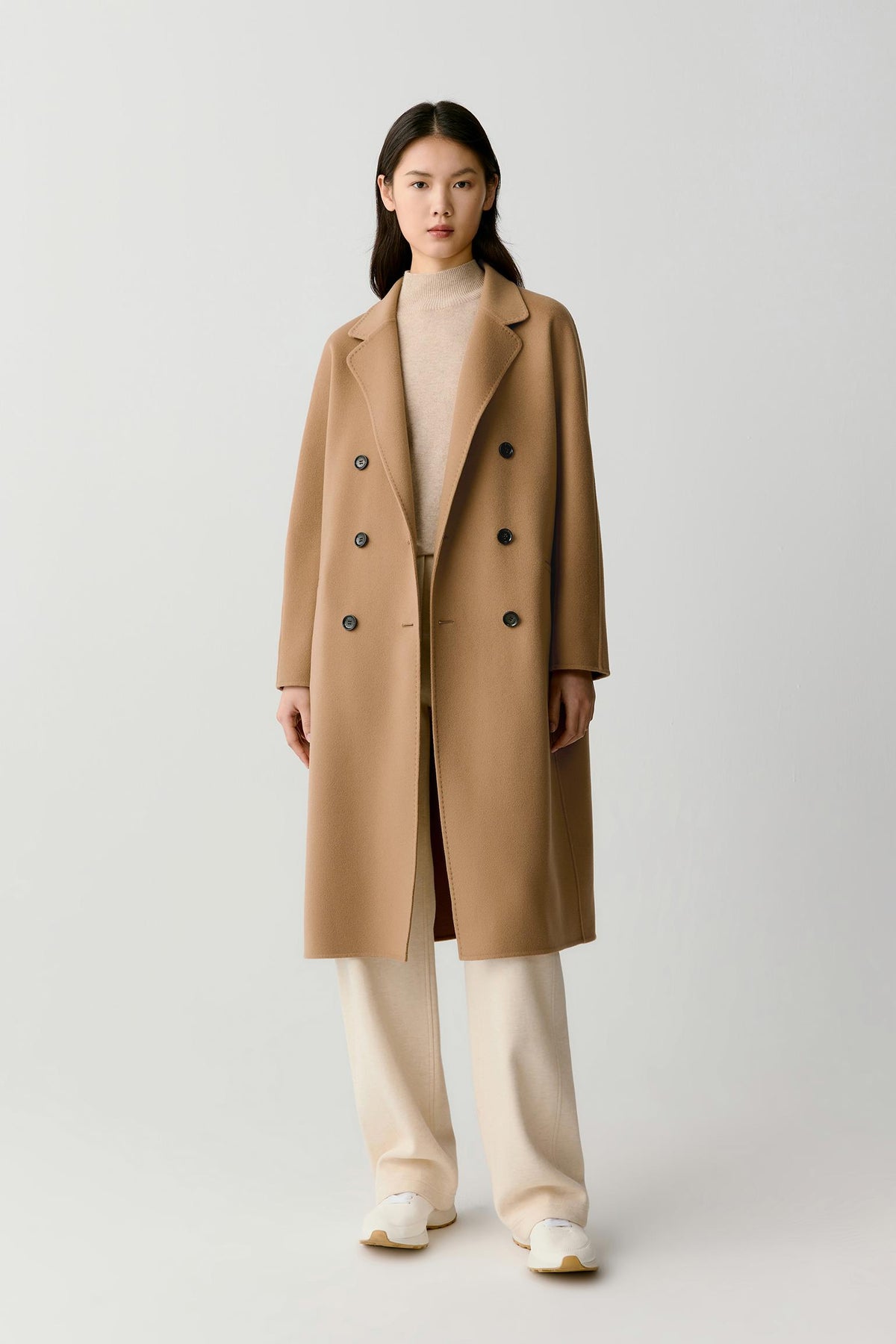 A model stands against a light gray studio background wearing a long camel double-breasted overcoat over a beige turtleneck, cream wide-leg trousers, and white sneakers.