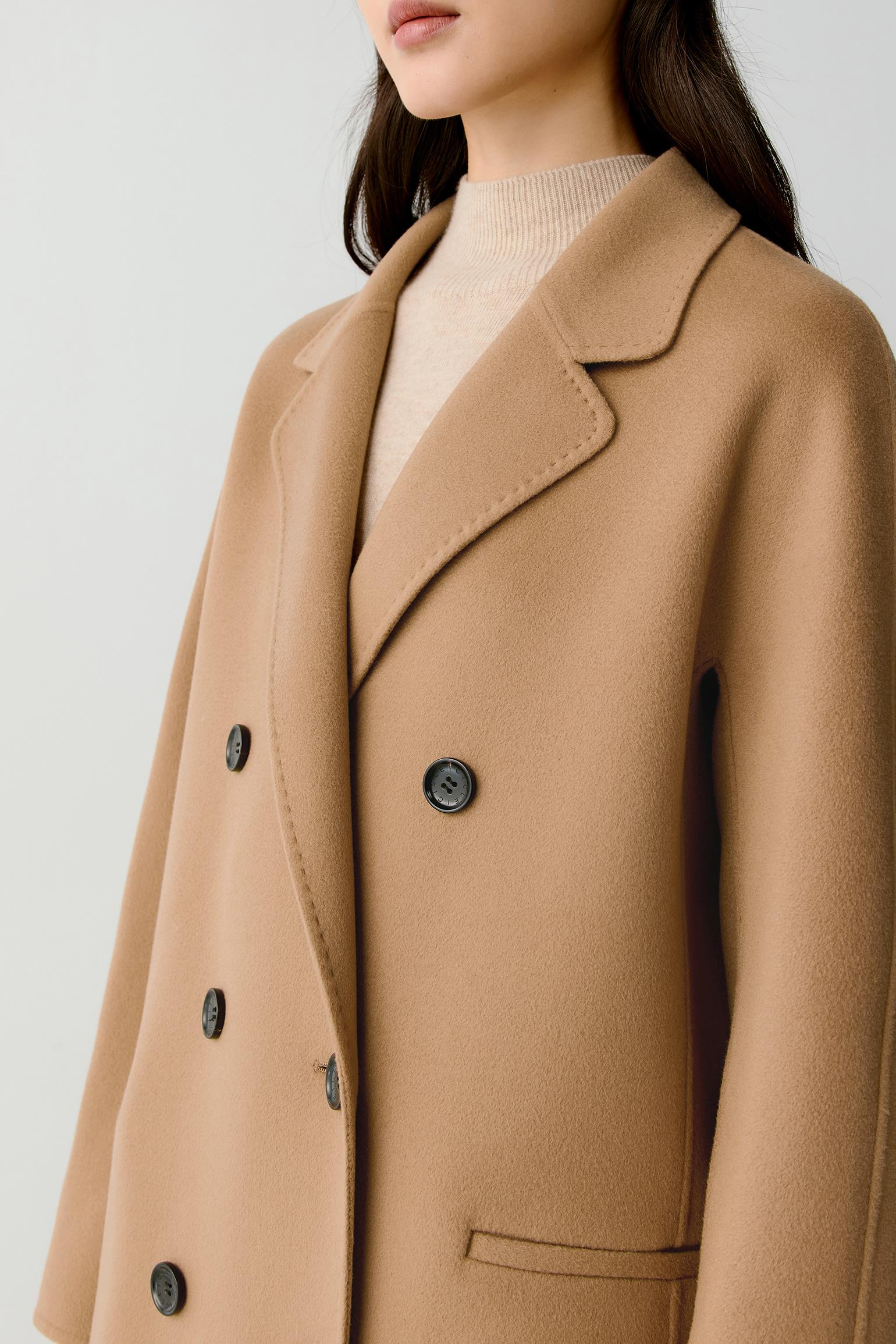 A close-up of a model wearing a camel-colored double-breasted wool coat over a cream ribbed turtleneck sweater, set against a plain light gray background.