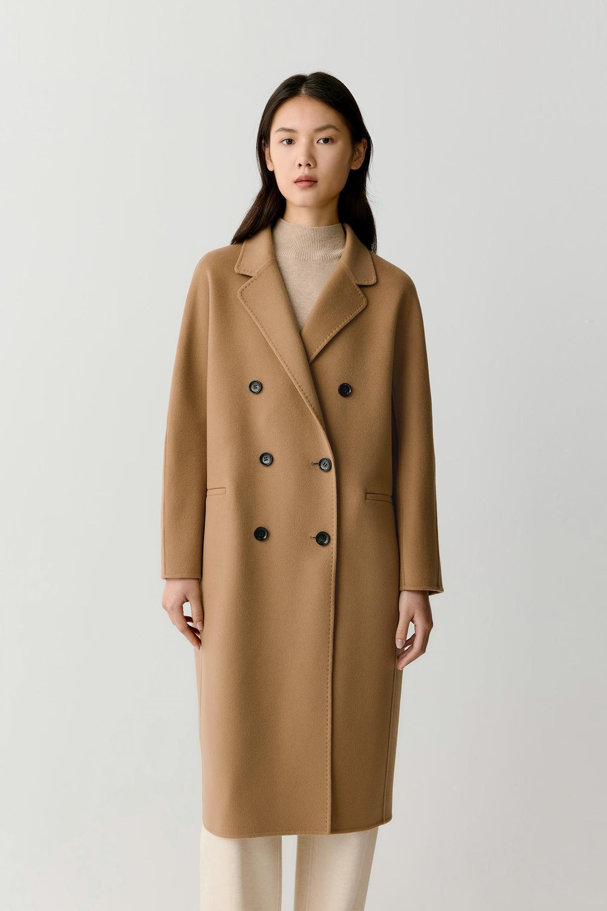 A woman stands against a plain light gray studio background wearing a camel-brown double-breasted coat over a beige top and light pants.
