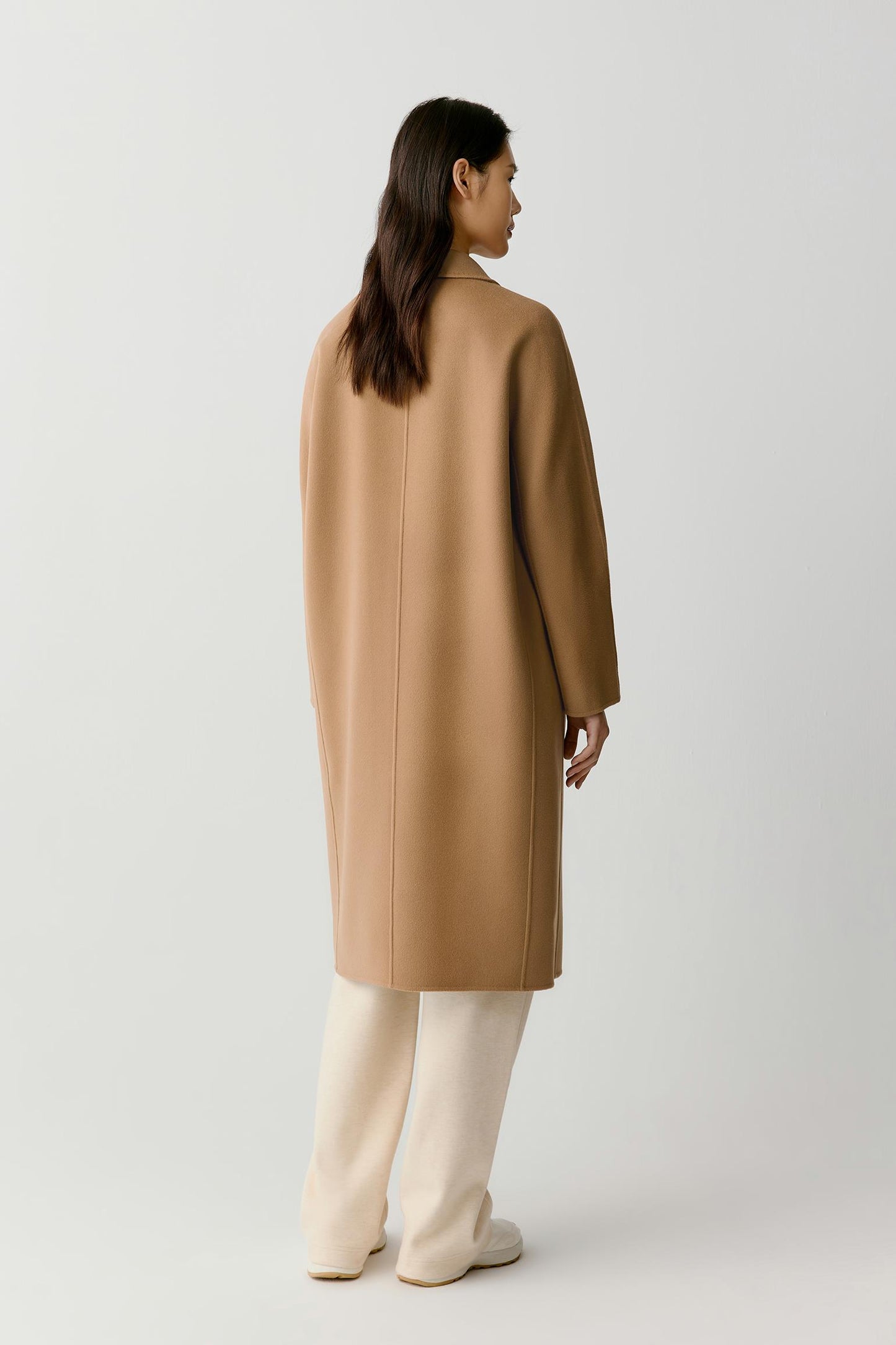 A woman shown from the back wearing a long camel-colored coat over cream wide-leg trousers, photographed against a clean light gray studio background.