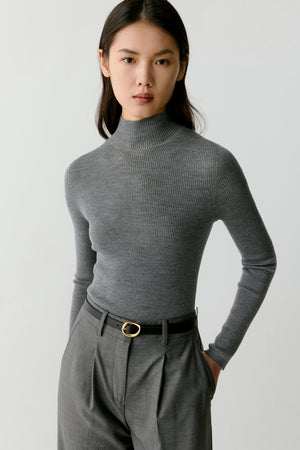 Front view of a model wearing a smoke grey ribbed pullover knitted from superfine wool. The sweater features a slim, body-skimming silhouette with a softly structured mock-neck collar that rises slightly above the neckline. The fine ribbed knit creates vertical texture across the entire garment while maintaining a smooth and lightweight surface. Long sleeves extend to the wrists and follow the natural shape of the arms. The pullover is styled with tailored grey trousers and a belt.