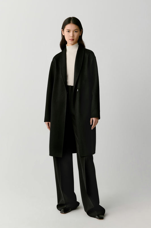 A model stands against a light gray studio background wearing a long black coat over a white turtleneck, black wide-leg pants, and black shoes.