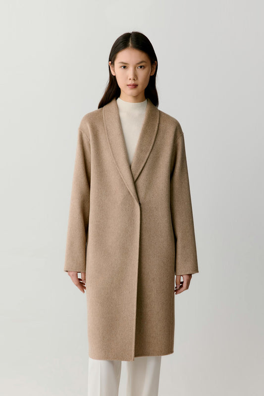 A woman modeling a long, open-front camel-colored coat with shawl-style lapels over a cream turtleneck and white trousers, photographed against a plain light gray studio background.