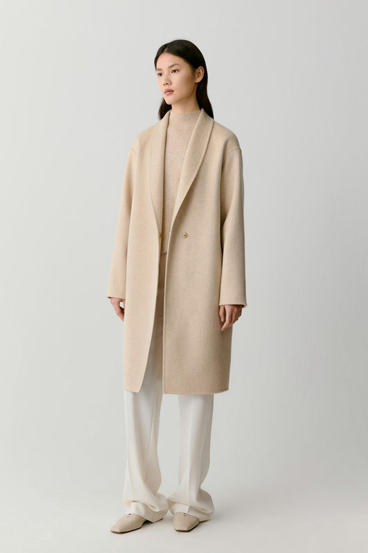 A model stands against a light gray studio background wearing a long, double-breasted beige wool-blend coat over a matching beige turtleneck, cream wide-leg trousers, and beige pointed flats.